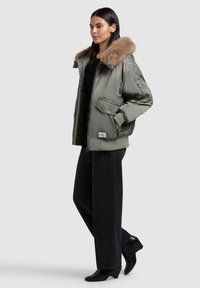 Giacca bomber verde oliva con colletto in pelliccia sintetica, dotata di tasche frontali, polsini a coste e chiusura con zip, abbinata a pantaloni neri a gamba larga.