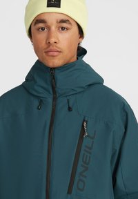 Veste teal avec une fermeture éclair à l'avant, capuche ajustable et logo noir. Présente une poche sur la poitrine avec un détail texturé et des fermetures éclair noires.