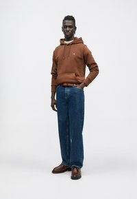 Barna kapucnis pulóver húzózsinórokkal, kék bő szárú farmernadrág, barna öv és sötét cipő. Egyszerű kialakítás apró logóval a kapucnis pulóveren.