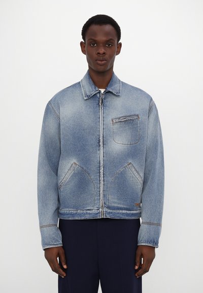 Hellblaues Denim-Jackett mit einem frontalen Reißverschluss, einer Brusttasche mit horizontalen Nähten und zwei geschwungenen Fronttaschen.