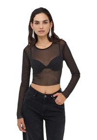 Top lungo a maniche nere in rete con pois bianchi e design corto, indossato sopra un reggiseno nero, abbinato a jeans neri a vita alta.