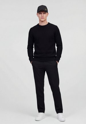 Uomo in piedi con le mani in tasca che indossa un berretto nero, un maglione nero, pantaloni neri e sneakers bianche su uno sfondo semplice.