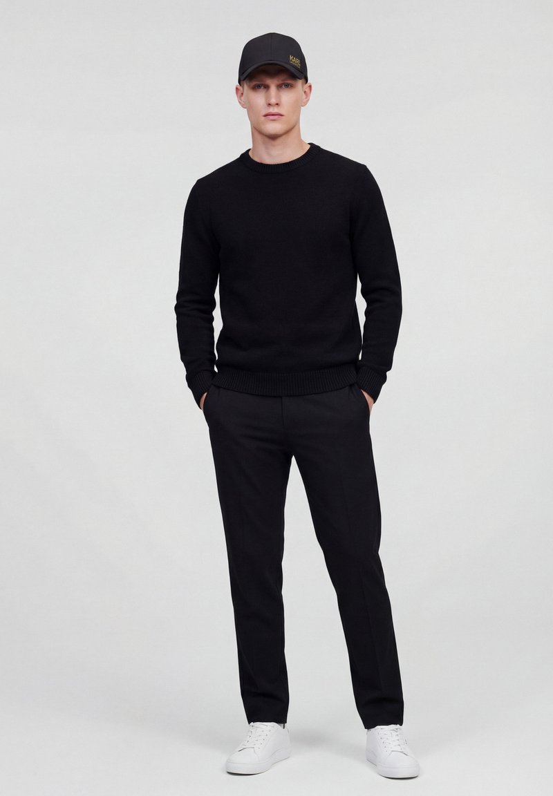 Homme debout, les mains dans les poches, portant une casquette noire, un pull noir, un pantalon noir et des baskets blanches, posé devant un fond uni.