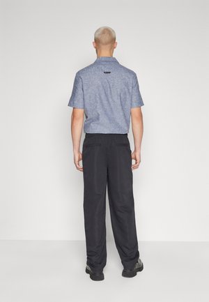 Camicetta blue in chambray a maniche corte con colletto, abbinata a pantaloni neri larghi. Sono indossate scarpe nere piatte.