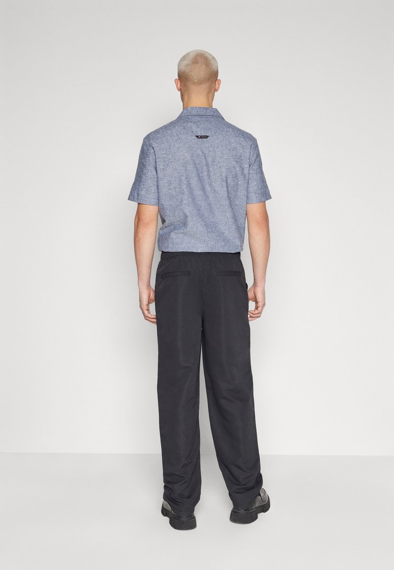 Kurzärmliges, blaues Chambray-Hemd mit Kragen, kombiniert mit lockeren, schwarzen Hosen. Es werden flache schwarze Schuhe getragen.