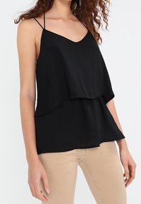 Top a canottiera nera a strati con spalline sottili, scollatura a V e tessuto leggero e fluido. Abbinato a pantaloni beige chiaro.