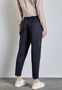 Un pantalon bleu marine présente une coupe slim, deux poches arrière et une poche latérale. Associé à des baskets blanches et un haut à manches longues beige.