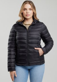 Vrouw draagt een zwart doorgestikt jack met capuchon, een gestreept shirt eronder en lichtblauwe spijkerbroek, staand tegen een effen achtergrond.