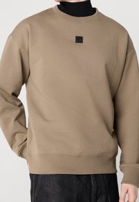 Uomo che indossa una felpa beige a collo rotondo sopra un dolcevita nero, con un logo quadrato nero sul petto, e pantaloni scuri, con una mano in tasca.