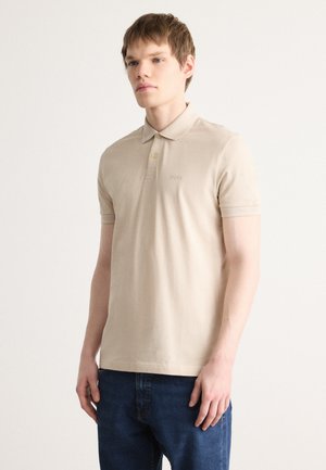 Polo beige en coton doux avec des manches courtes, un col à boutons et des détails de logo discrets sur la poitrine. Associé à un jean en denim bleu.