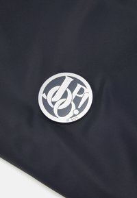 Námornícka látka s hladkým povrchom, ktorá obsahuje okrúhly kovový logo v striebornej farbe s textom "JP! JEANS" na spodnej strane, zdôrazňujúca detaily dizajnu.