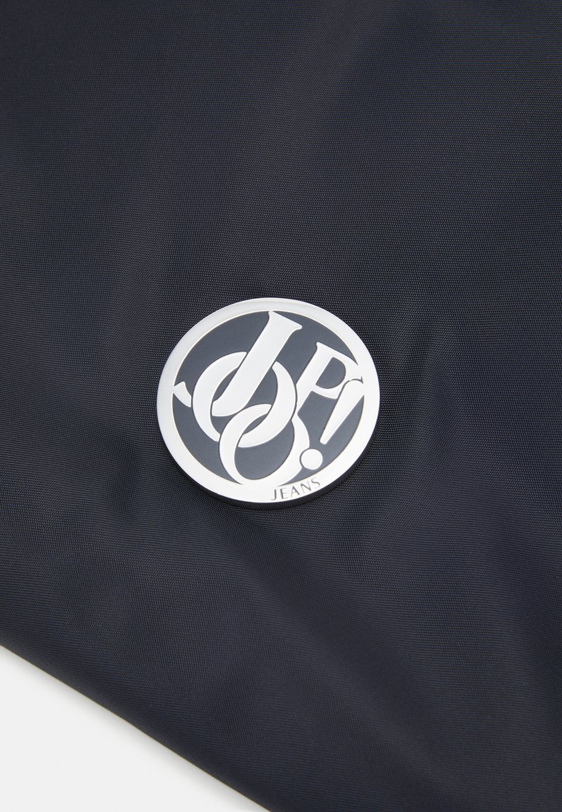 Marineblaues Gewebe mit glatter Textur, das ein rundes metallisches Logo in Silber mit dem Text "JP! JEANS" am unteren Rand zeigt und damit das Design betont.