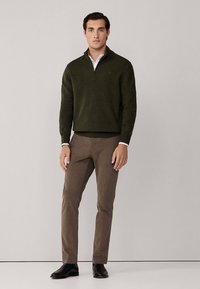 Sötétzöld, bordázott textúrájú cipzáros pulóver, bézs chino nadrággal és fekete cipővel párosítva. A modell világosszürke háttér előtt áll.