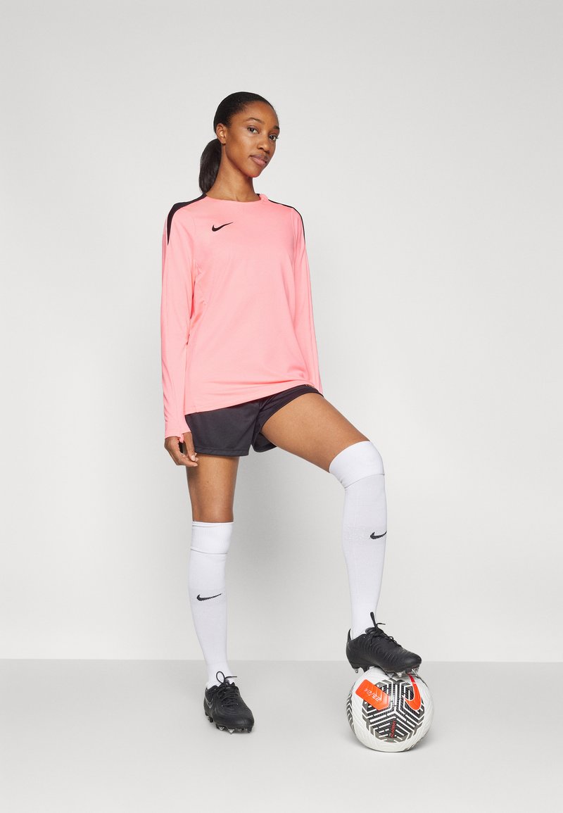 Maillot long à manches Nike rose avec des accents noirs, associé à un short gris, des chaussettes blanches montantes et des crampons noirs, se tient sur un ballon de football à motifs.