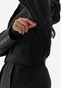 Černá výkonnostní bunda s hladkou texturou, přiléhavým střihem, kapsou na zip a nastavitelným pasem se značkovým detailem.