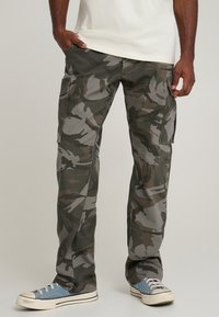 Cargo pants in camo print featuring shades of green and gray, with multiple side pockets and a relaxed fit, paired with blue sneakers.