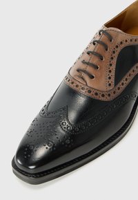 Černá kožená bota s špičatou špičkou, hnědým koženým akcentem s perforovanými detaily a hnědými tkaničkami. Obsahuje brogue vzory a hladký povrch.