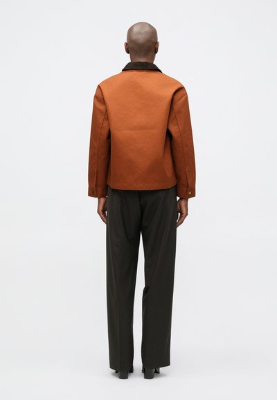 Barna, strukturált zakó gallérral, hátul redőkkel és gombos mandzsettával, sötét nadrággal és magassarkú cipővel párosítva, hátulnézetből.