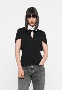 Schwarzes Oberteil mit einem capeartigen Design und einem weißen Kragen. Besitzt ein Schlitzdetail vorne. Kombiniert mit grauen Jeans.
