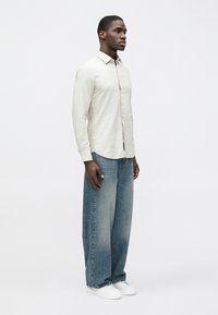 Svetlobéžová košeľa s dlhými rukávmi a zapínaním na gombíky, kombinovaná s voľnými vyblednutými džínsami a bielymi teniskami. Neformálny, uvoľnený strih.