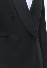 Μαύρο σακάκι διπλής σειράς με κουμπιά καλυμμένα με ύφασμα και στενές πέτες, φορεμένο πάνω από λευκό πουκάμισο ή μπλούζα.