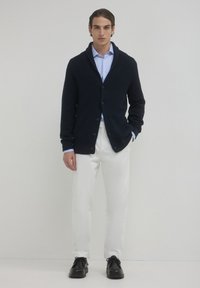 Uomo che indossa un cardigan strutturato blu navy sopra una camicia azzurra, pantaloni bianchi e scarpe stringate in pelle nera, in piedi davanti a uno sfondo bianco neutro.