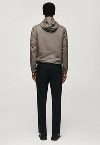 Hellbraune Kapuzenjacke mit elastischen Bündchen, Rückennahtdetail, kombiniert mit schwarzen Hosen und schwarzen Schuhen, von hinten betrachtet.