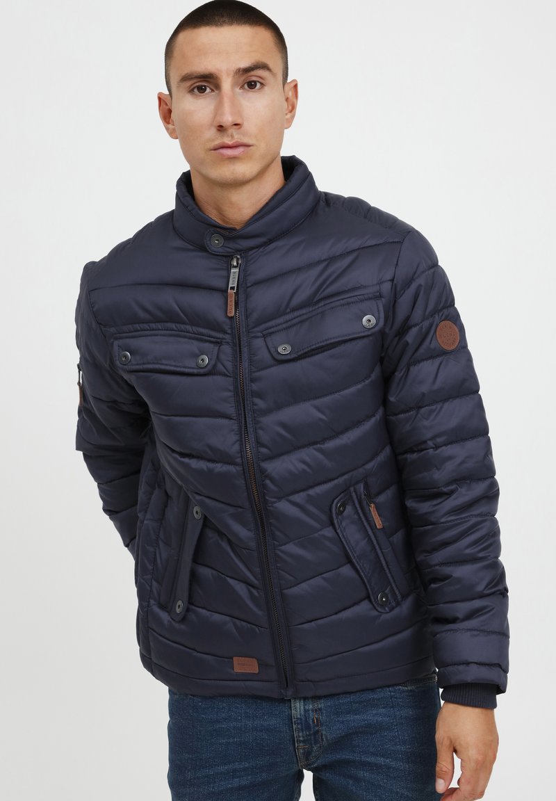 Navy puffer jacket with a high collar, horizontal quilted pattern, two chest pockets, side pockets, and leather branding detail on the sleeve.