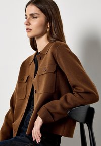 Bruine gewaxt jasje met een kraag, twee voorzakken en bruine knopen. Draagt over een zwarte top, met een gladde, aansluitende silhoeet.