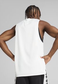 Camiseta atlética sin mangas blanca de tejido transpirable. Presenta un corte relajado, cuello redondo y una textura suave, llevada sobre pantalones cortos negros.