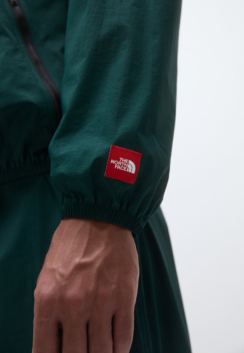 Grüne, leichte Jacke mit elastischen Bündchen, ausgestattet mit einem roten Logo-Patch von The North Face am Ärmel und einem Reißverschluss-Design auf der Vorderseite.