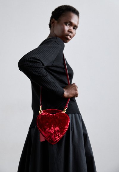 Sac en velours rouge en forme de cœur avec une longue sangle et des accessoires en or, porté avec un pull noir en maille côtelée et une jupe noire fluide.