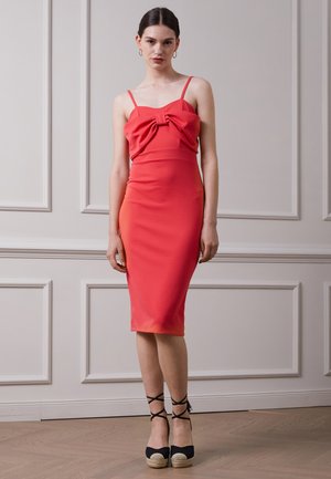 Vestido coral ajustado com um detalhe de laço no busto, apresentando alças ajustáveis e uma saia até ao joelho. Sandálias de espadrilhe com atilhos para os tornozelos.