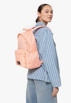 Mochila de color melocotón con acabado suave, correas ajustables, bolsillo frontal de malla y cierre de cordón, llevada sobre una camisa de rayas azules.