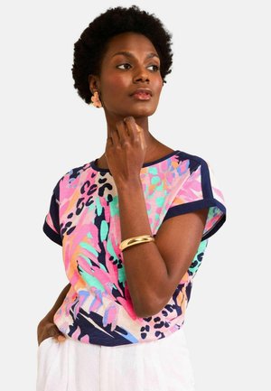 Woman with short curly hair wearing a colorful abstract print top, gold bracelet, flower-shaped earrings, and light-colored pants.