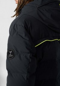 Zwarte donsjas met capuchon, met een ronde JOTT-logo op de linkerarm en een neongele accent langs de schoudernaad.