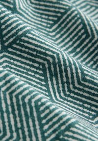 Detailný záber na modrozelenú a bielu látku s geometrickým labyrintovým vzorom v tvare cikcak a viditeľnými záhybmi textúry.