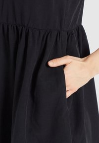 Main placée dans la poche latérale froncée d'une robe noire, montrant la texture du tissu et les détails du design de la poche.