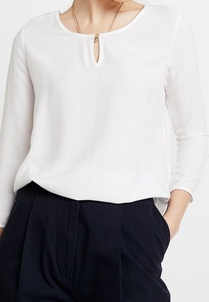 Kobieta nosząca białą bluzkę z długim rękawem, z małym otworem na przodzie i złotym zapięciem, w połączeniu z czarnymi spodniami z wysokim stanem.