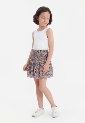 Dziewczyna z krótkimi brązowymi włosami, ubrana w biały bezrękawnik, szarą warstwową spódnicę w panterkę i białe trampki, stoi na białym tle.