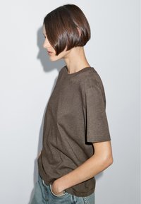 T-shirt marrone a maniche corte in tessuto morbido, con scollatura rotonda e vestibilità rilassata, indossato con jeans azzurri chiari.