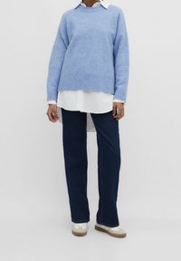 Hellblauer Strickpullover mit Rippabschluss, über einem weißen Hemd mit Kragen getragen. Kombiniert mit dunkelblauen Straight-Leg-Jeans und weißen Sneakers.
