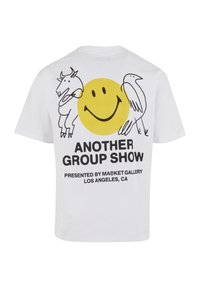 Hvit bomull t-skjorte med et grafisk motiv av et gult smilende ansikt med en ku og en fugl, samt tekst som sier "EN ANNEN GRUPPEUTSTILLING."