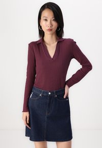 Burgundska rebrasta majica z dolgimi rokavi in V-izrezom ter temno jeans mini krilo v A-liniji, ki ima žep in gumbasto zapiranje. Gladke teksture ustvarjajo kontrast.
