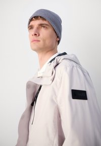 Light-colored, waterproof jacket with a hood, featuring black accents and a logo patch. Worn with a textured gray beanie.