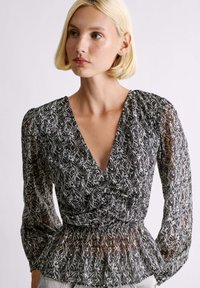 Čierno-biela blúzka s vzorom, hlbokým výstrihom do V, dlhými nafúknutými rukávmi a naškrobeným pásom, vyrobená z ľahkej, textúrovanej látky.