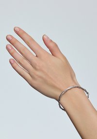 Srebrna bransoletka o teksturowanym, plecionym wzorze noszona na dłoni. Bransoletka ma gładkie krawędzie i delikatny styl.