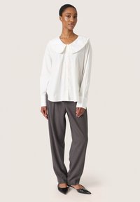 Camicia bianca con colletto ampio e volant, abbinata a pantaloni grigi a righe sottili e scarpe nere a punta. Tessuto morbido.