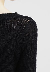 Schwarzer gestrickter Pullover mit lockerem Gewebe, der einen gerippten Halsausschnitt und ein strukturiertes Muster auf der Rückseite zeigt. Weicher Stoff mit entspanntem Sitz.