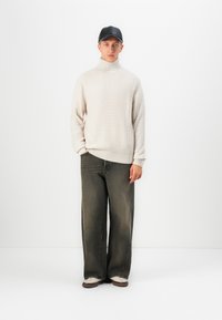 Young man standing wearing beige turtleneck sweater, black baseball cap, faded wide-leg jeans, and white sneakers against a plain white background.